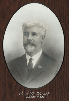 Black and white photograph of an oval portrait of a suited man with thick white hair and goatee.
