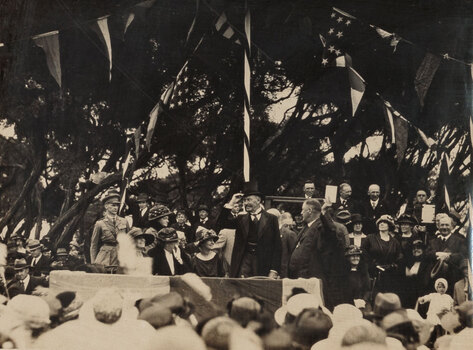 Sepia photograph of a man in a top hat upon a stage in front of a large crowd of people