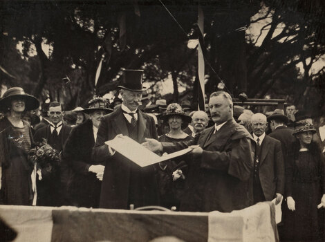 Sepia photograph of a man in a top hat upon a stage being presented an illuminated certificate in front of a crowd of people