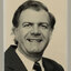 Black and white photographic portrait of a man in a dark suit and tie 