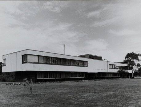 Black and white photograph of a modern office building, 2 stories high, with grass in front and clouds above.