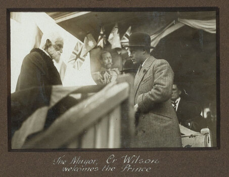Black and white photograph of a man with white hair and a beard shaking hands with a man in a bowler hat. Behind them are flags