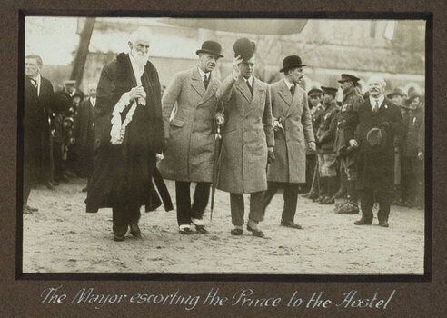 Black and white photograph of a man with white hair in mayoral robes standing next to 3 men with bowler hats, man in centre has his raised.