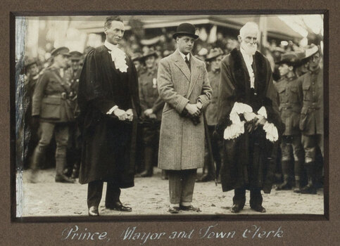 Black and white photograph of three men, men on left (Town clerk) and right (Mayor) wear ceremonial robes, man in centre (The Prince) is in bowling hat