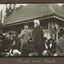 Black and white photograph of 2 men in front of a small building, surrounded by crowd.
