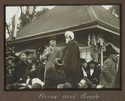 Black and white photograph of 2 men in front of a small building, surrounded by crowd.