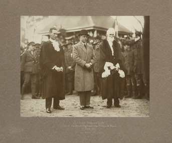 Sepia photograph of three men standing in foreground, man in centre in bowler had next to two men in ceremonial robes. Uniformed men in the background in front of a building