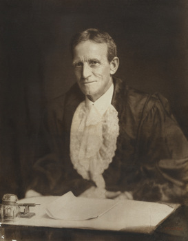 Formal portrait of seated man wearing mayoral robes with fur trims and lace cuffs and jabot, he sits at a writing desk. He has short dark hair and is clean shaven.