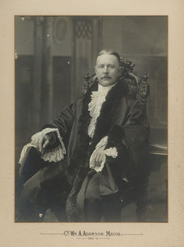 A formal portrait of seated man wearing mayoral robes with fur trims and lace cuffs and jabot. He sits in a decoratively carved chair. He has short hair and a moustache. 