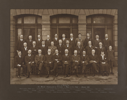 Sepia photograph of Mayor, Councillors and Officers seated and standing in front of the Brighton Town Hall. 31 people total, of which one is a boy and one is a woman. 
