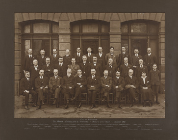 Sepia photograph of Mayor, Councillors and Officers seated and standing in front of the Brighton Town Hall. 31 people total, of which one is a boy and one is a woman. 