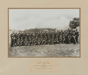 Four rows of uniformed men, two rows standing, one row kneeling and one row sitting. Taken in an outdoor setting with tents in background