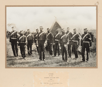 Group of eleven men in dark uniform with white sash across torso and wearing hats standing on grass. Some men are holding swords. Tents and more soldiers in background