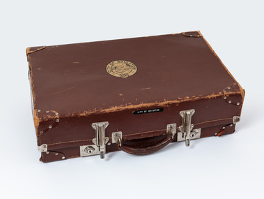 Brown leather suitcase, worn around the edges, with metal clasps and leather handle. Has a gold sticker on top bearing Brighton Coat of Arms. 