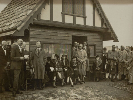 Black and white photograph of a suited man delivering a speech to an audience in front of a small building
