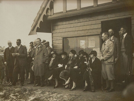 Black and white photograph of a suited man giving a speech to a crowd outside by a small building