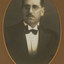 Black and white photograph of a man with dark hair and glasses wearing a dark suit and bow tie