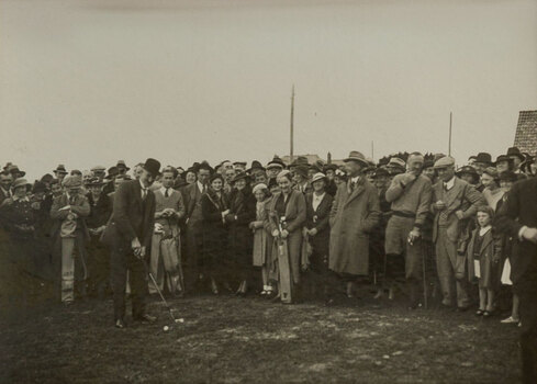 Black and white photograph of a suited man hitting a golf ball in front of a crowd