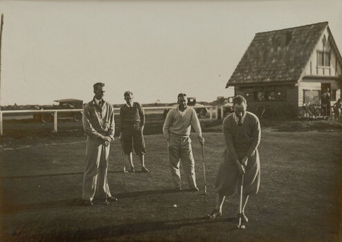 Black and white photograph of four men playing golf, a small building in the background