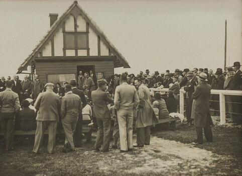 Black and white photograph of a crowd gathering around officials outside of a small building