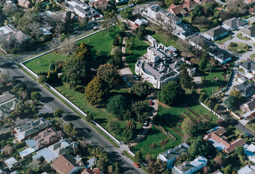 Aerial photograph of large white mansion set within verdant grounds. Streets and other buildings surrounding the gardens. 
