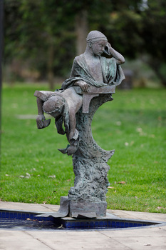 Outdoor sculpture of two children above a trunk, below them is a half circle filled with water. Set within green parklands behind