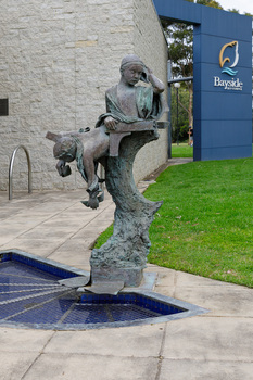 Outdoor sculpture of two children above a trunk, below them is a half circle filled with water. Behind the sculpture is the Bayside City Council office building with grass to the right