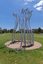 Photograph of a silver sculpture that look like large reeds in a semi-circular shape with a bench sear within. The sculpture is set within a green park.