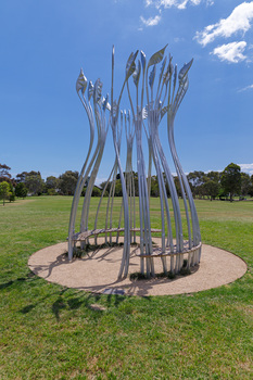 Photograph of a silver sculpture that look like large reeds in a semi-circular shape with a bench sear within. The sculpture is set within a green park.