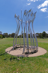 Photograph of a silver sculpture that look like large reeds in a semi-circular shape with a bench sear within. The sculpture is set within a green park.