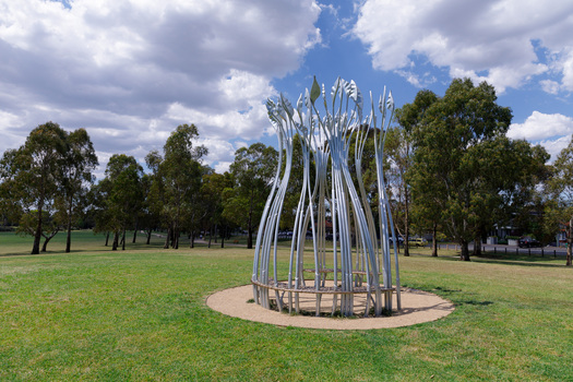 Photograph of a silver sculpture that look like large reeds in a semi-circular shape with a bench sear within. The sculpture is set within a green park.