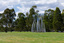 Photograph of a silver sculpture that look like large reeds in a semi-circular shape with a bench sear within. The sculpture is set within a green park.