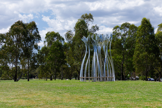 Photograph of a silver sculpture that look like large reeds in a semi-circular shape with a bench sear within. The sculpture is set within a green park.