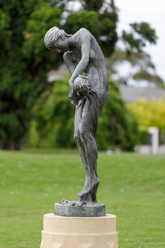 Bonze sculpture of a classical female figure who holds a vessel and looks downward. The sculpture sits on a concrete pedestal surrounded by greenery.