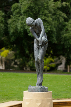 Bonze sculpture of a classical female figure who holds a vessel and looks downward. The sculpture sits on a concrete pedestal within a pond, surrounded by greenery.
