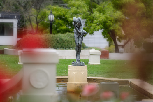 Bonze sculpture of a classical female figure who holds a vessel and looks downward. The sculpture sits on a concrete pedestal within a pond, surrounded by greenery.