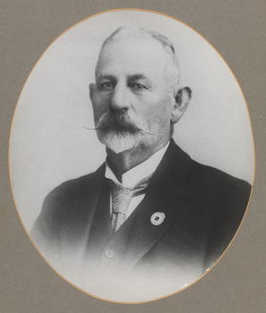 Black and white oval shaped photograph of a formal portrait of a man, from chest up wearing a dark suit.