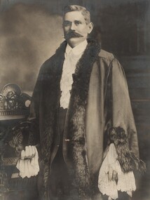 Formal portrait of a man with dark hair and a thick moustache standing, wearing mayoral robes with fur trims, lace cuffs, jabot and white shirt underneath.