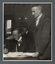 Black and white photograph of a man seated at a desk with a second man standing next to him