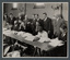 Black and white photograph of people seated at a long trestle table with piles of documents in front of them, a line of people stand behind