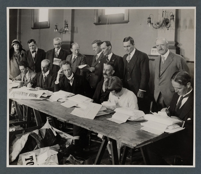 Black and white photograph of people seated at a long trestle table with piles of documents in front of them, a line of people stand behind
