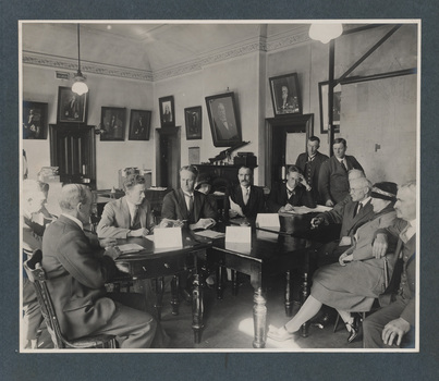 Black and white photograph of suited men and two women surrounding a curved desk and looking at documents