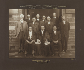 Black and white photograph of suited men in three rows in front of an official building, one man is wearing mayoral robes. The photograph sits on a dark mount with handwritten inscriptions above and below