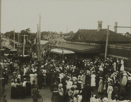Black and white photograph of a large crowd around a tram by a train station