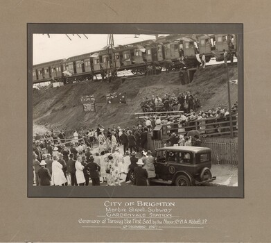Black and white photograph of a speech being delivered by officials on a platform on a railway embankment in front of a gathered crowd. At the top of the embankment behind them is a train, and there are flags and buntings hung as decorations. 