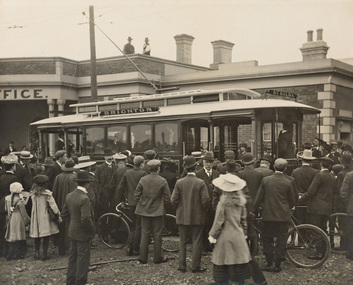 Black and white photograph of the opening of the St Kilda-Brighton Electric Street tram, surrounded by a crowd of people in the foreground, and a building in the background. 