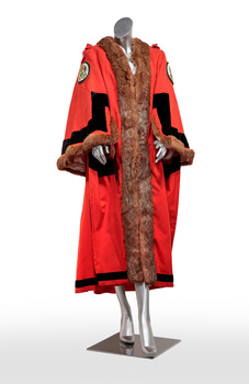Photograph of a ceremonial robe on a silver mannequin. The robe is red with brown fur trim and black lines. It has a Coat of Arms embroidered on the shoulders.