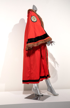 Photograph of a ceremonial robe on a silver mannequin taken on the side. The robe is red with brown fur trim and black lines, it has coat of arms embroidered on the shoulders