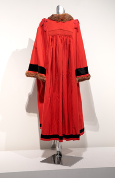 Photograph of a ceremonial robe on a silver mannequin taken from the back. The robe is red with brown fur trim and black lines.