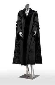Photograph of a full length black robe with centre opening on a silver mannequin. Has black velvet edging and is adorned with tassels.